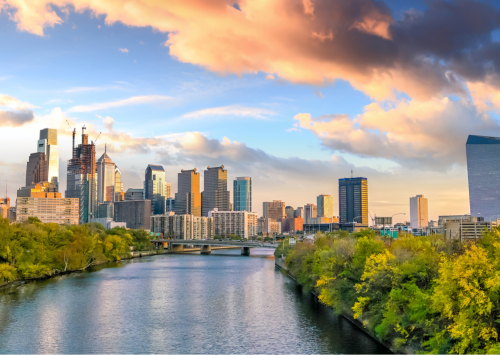 Philadelphia skyline at sunset with skyscrapers, a river, trees, and colorful clouds in the sky.